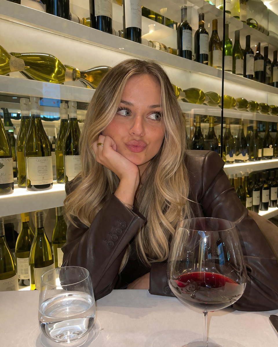 Candid Wine Bar Portrait のQwen Imageプロンプト例: Generates a photorealistic, candid-style portrait of a person sitting at a table with drinks and a background of illuminated shelves.