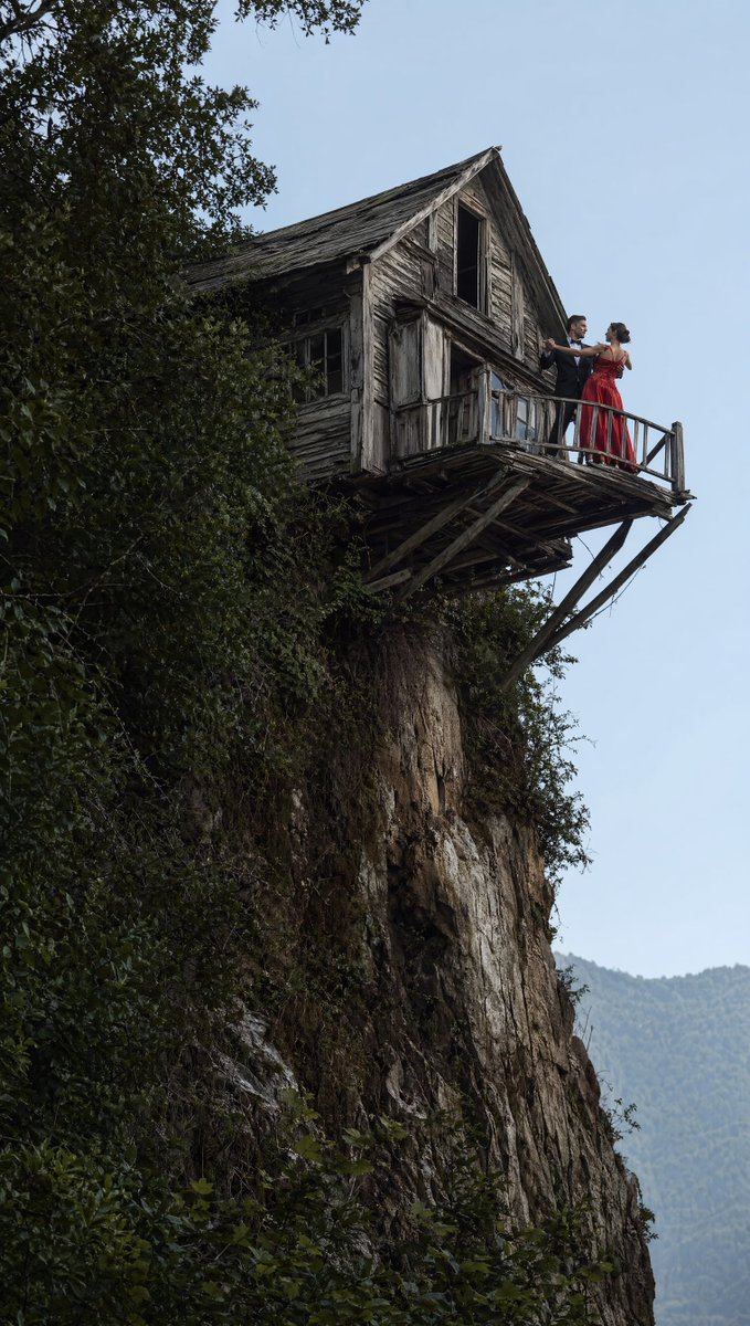Dancing Couple on Cliffside House のQwen Imageプロンプト例: A moody cinematic photo prompt of an elegant couple dancing on the balcony of a decaying house perched dangerously on a sheer...