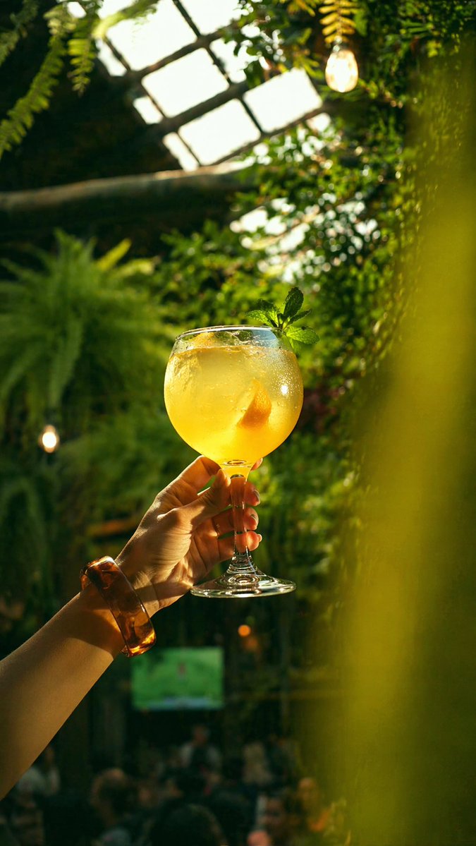 Golden Cocktail in Greenhouse Bar Qwen Image prompt example: This prompt generates a photorealistic lifestyle shot of a hand raising a glowing citrus cocktail in a lush...