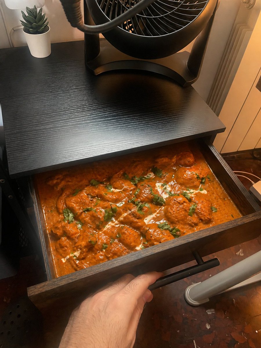 Chicken Tikka Masala Mug Desk Photo のQwen Imageプロンプト例 バリエーション 3: A photorealistic joke snapshot of a hand holding a black mug filled with chicken tikka masala in a warm-lit desk...