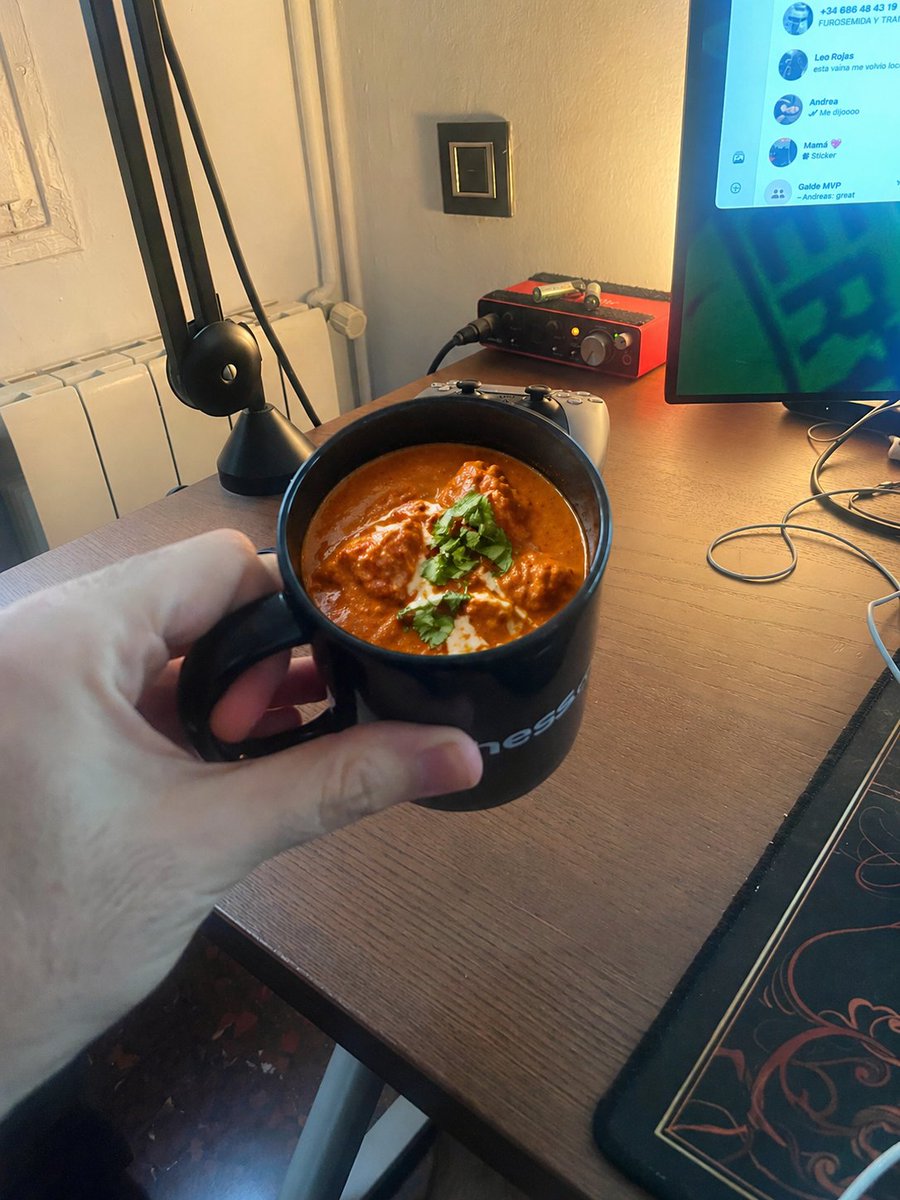 Chicken Tikka Masala Mug Desk Photo のQwen Imageプロンプト例: A photorealistic joke snapshot of a hand holding a black mug filled with chicken tikka masala in a warm-lit desk setup,...